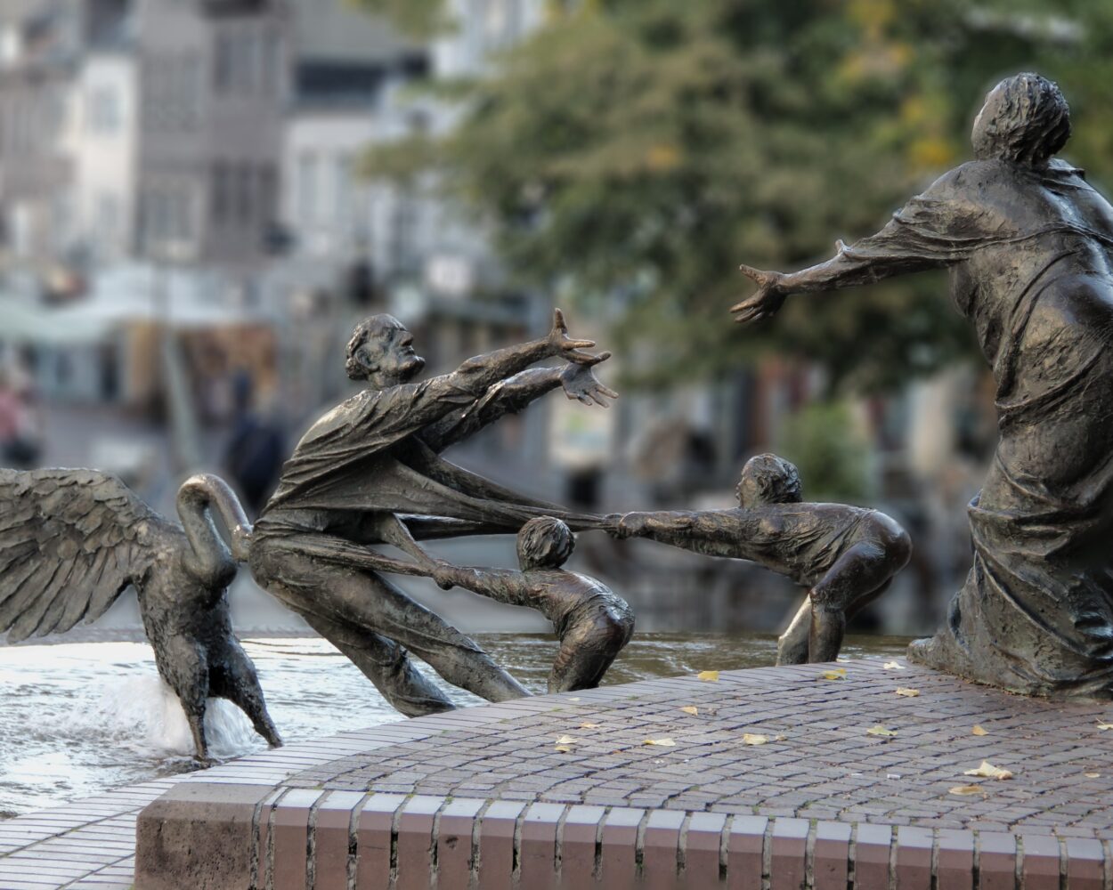 Bronzeskulpturengruppe im Elsabrunnen mit einer Frau, die zwei Kinder an den Händen hält, während ein Engel sie umarmt. Im Hintergrund sind Cafés und Bäume sichtbar.