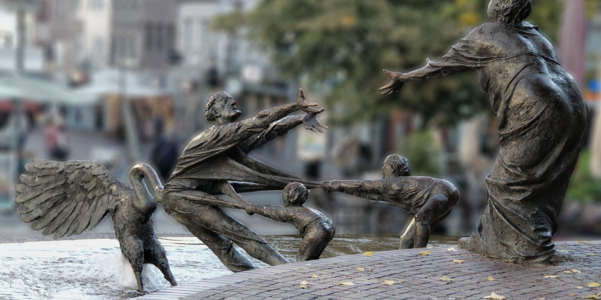 Bronzeskulpturengruppe im Elsabrunnen mit einer Frau, die zwei Kinder an den Händen hält, während ein Engel sie umarmt. Im Hintergrund sind Cafés und Bäume sichtbar.