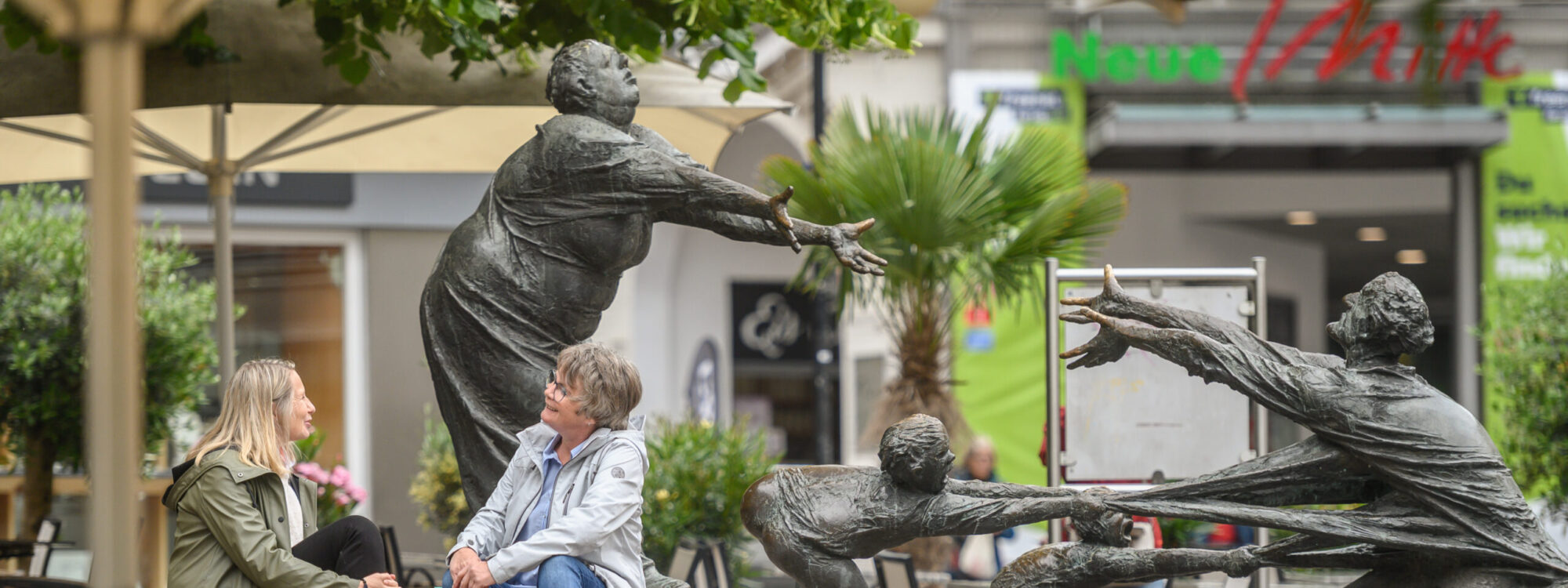 Elsabrunnen im Hintergrund, im Vordergrund Gäste in einem Café.
