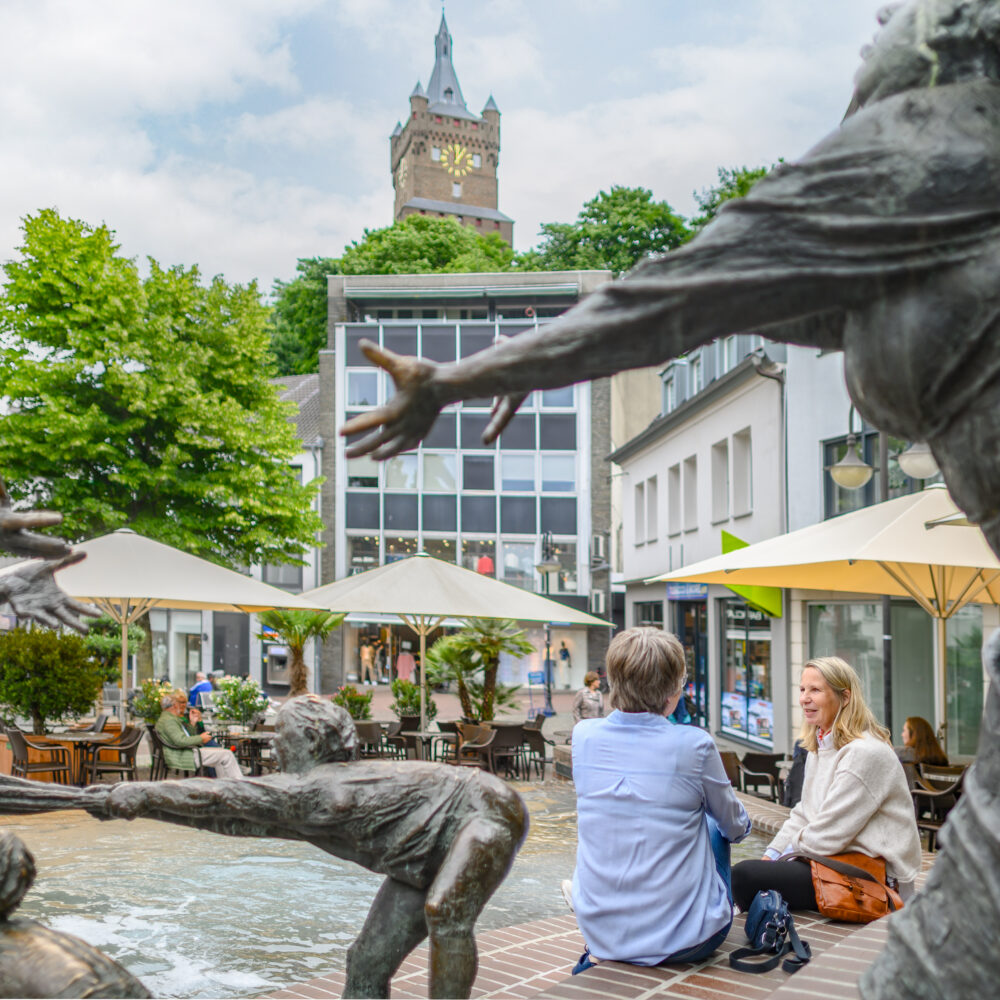 Bronze-Skulpturen von Menschen, die aus einem Brunnen herausragen, im Vordergrund. Im Hintergrund sitzen zwei Personen auf einer Bank, umgeben von Bäumen und Cafétischen mit Sonnenschirmen. Die Schwanenburg ist sichtbar.