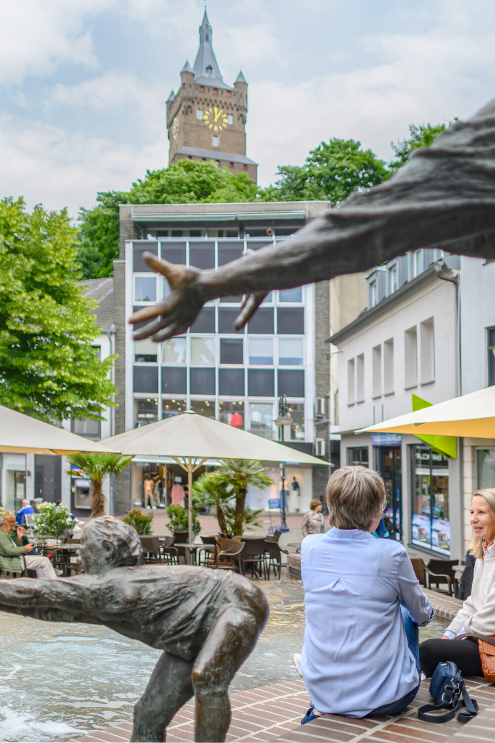 Bronze-Skulpturen von Menschen, die aus einem Brunnen herausragen, im Vordergrund. Im Hintergrund sitzen zwei Personen auf einer Bank, umgeben von Bäumen und Cafétischen mit Sonnenschirmen. Die Schwanenburg ist sichtbar.