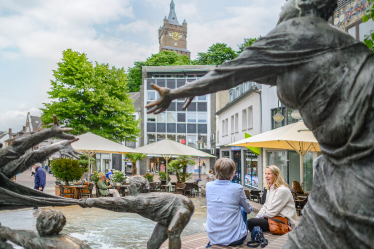 Bronze-Skulpturen von Menschen, die aus einem Brunnen herausragen, im Vordergrund. Im Hintergrund sitzen zwei Personen auf einer Bank, umgeben von Bäumen und Cafétischen mit Sonnenschirmen. Die Schwanenburg ist sichtbar.