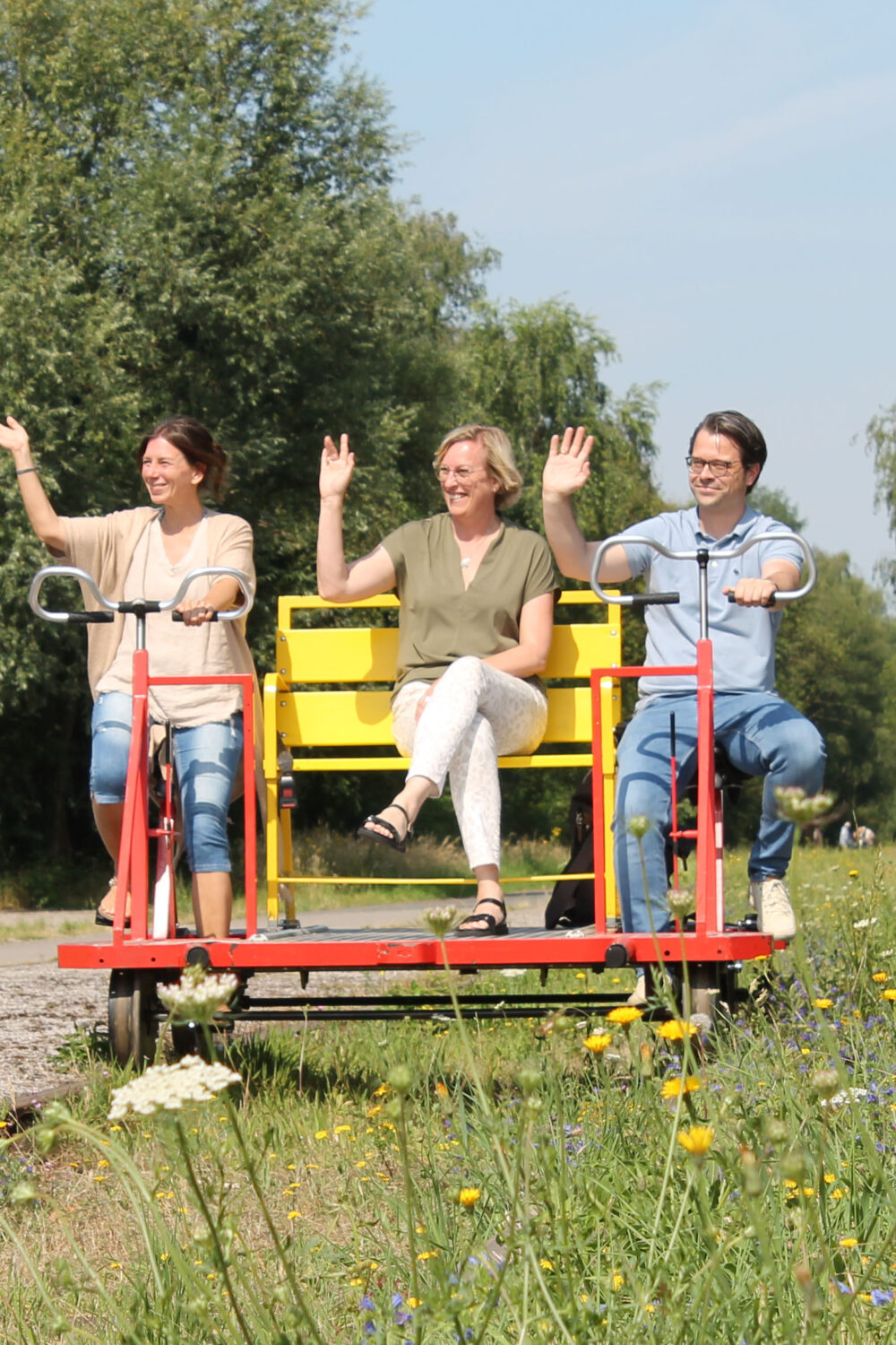 Drei Personen sitzen auf einer roten Fahrraddraisine mit gelben Sitzen von der Grenzland-Draisine. Sie winken in die Kamera. Im Hintergrund sind Bäume und eine Uhr zu sehen.