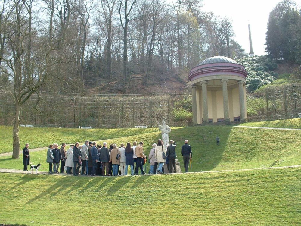 Gruppe von etwa 20 Personen bei einer Führung durch die Gartenanlagen vor einem runden Pavillon mit Säulen. Im Hintergrund ist eine Statue sichtbar