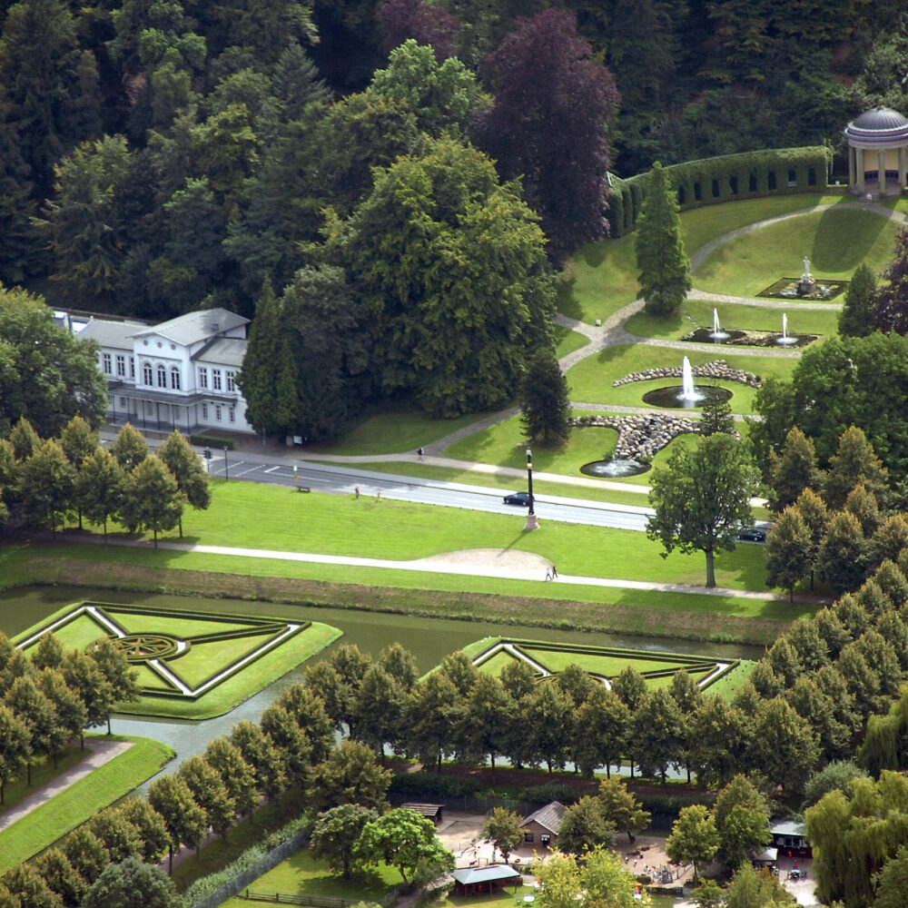 Luftaufnahme der symmetrisch gestalteten Klever Gartenanlage mit Hecken, einem Springbrunnen, einem kleinen Pavillon und von Bäumen umgebenen Wegen.