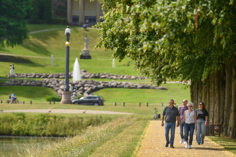 Vier Spaziergänger gehen auf einem Weg entlang eines Parks mit Bäumen. Im Hintergrund ist ein Brunnen und eine Statue sichtbar.