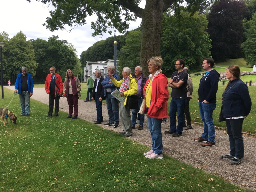 Gruppe von elf Personen steht auf einem Weg in den historischen Gartenanlagen, umgeben von Bäumen und Rasen, eine Person hält eine Karte oder ein Buch.