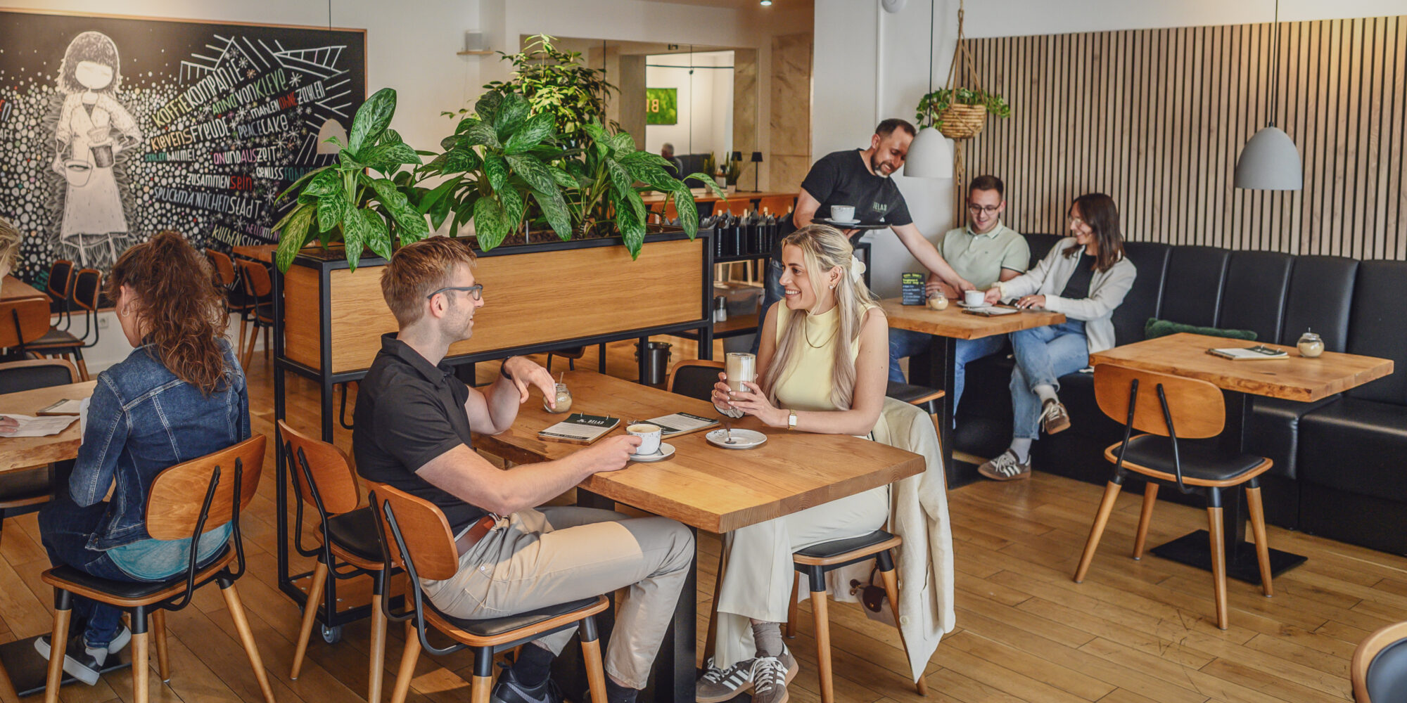 Innenansicht des Kavariner Cafés mit mehreren Tischen aus Holz. Gäste sitzen auf Stühlen, einige trinken Kaffee. Pflanzen dekorieren den Raum.