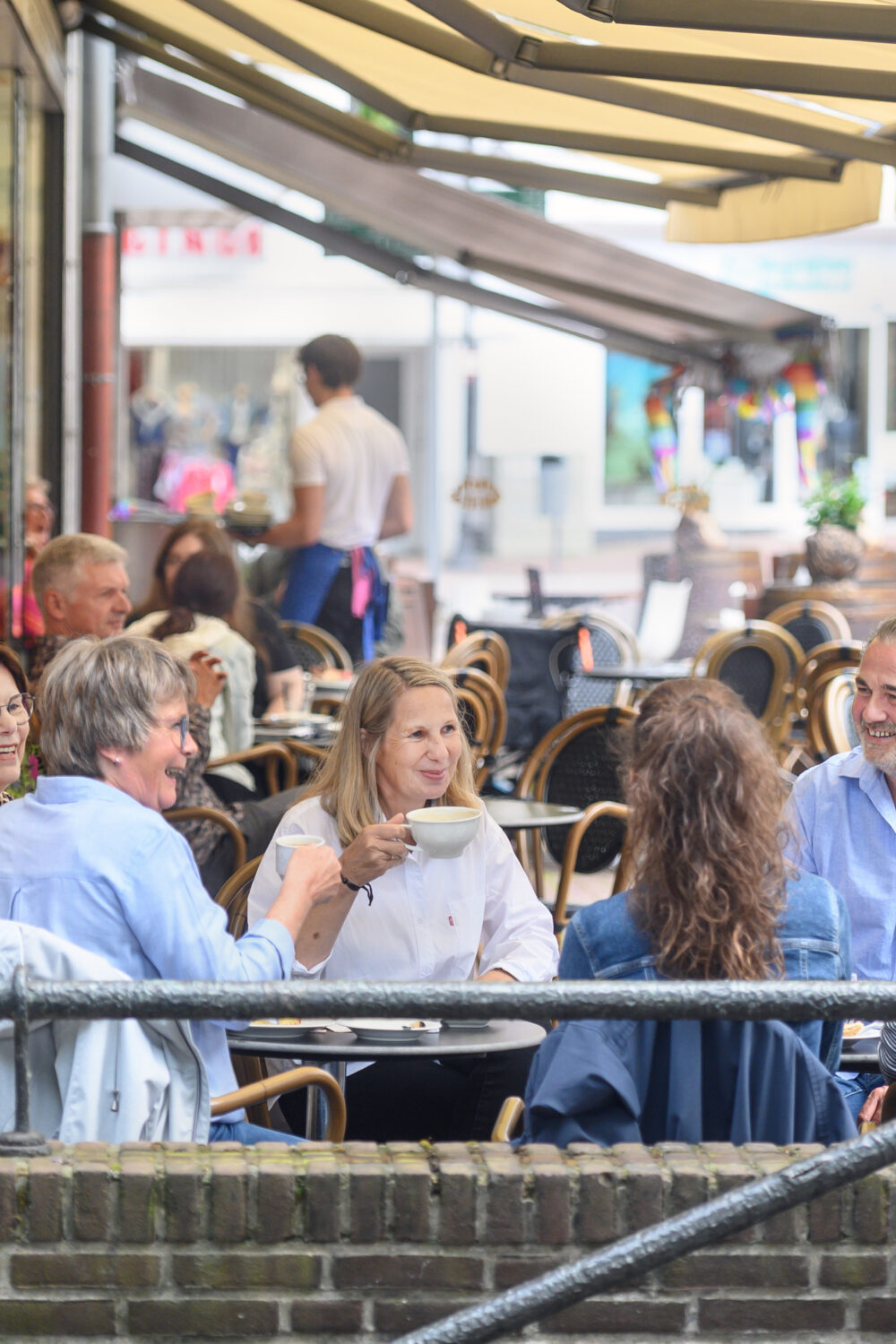 Sechs Personen sitzen in Kleve an zwei Tischen im Außenbereich eines Cafés mit braunen Stühlen und Tischen, umgeben von Bäumen und Gebäuden.