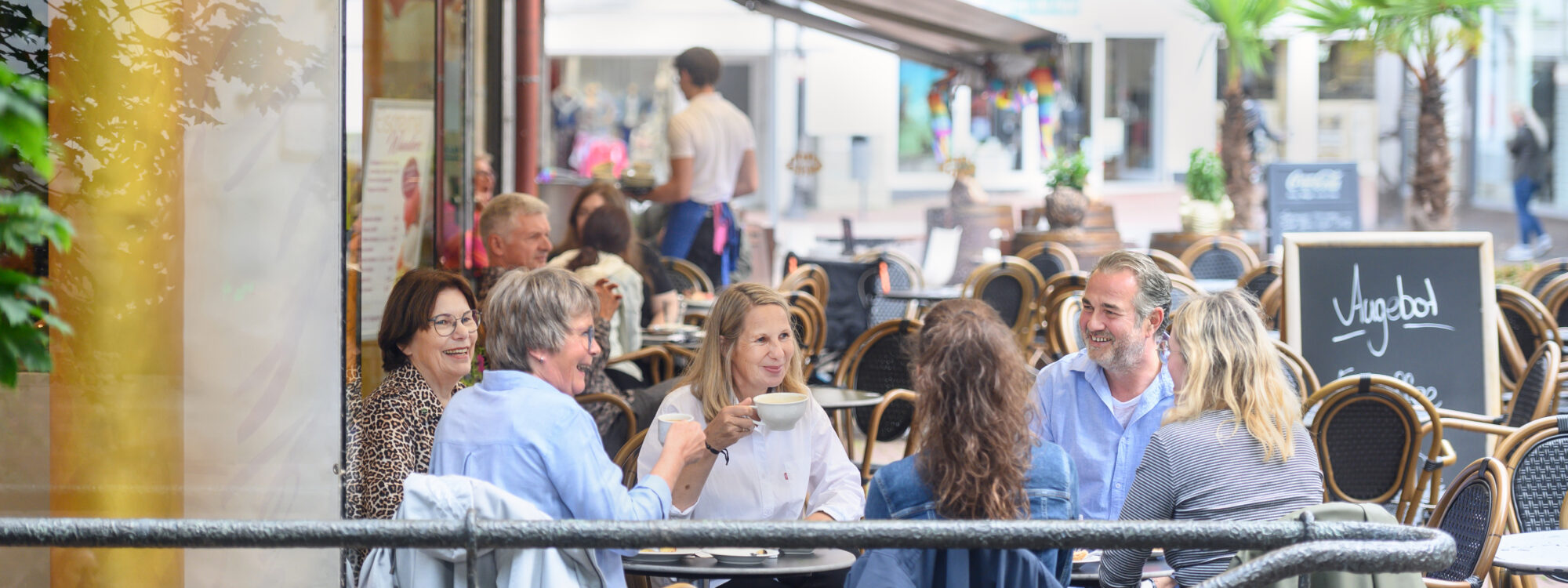 Sechs Personen sitzen in Kleve an zwei Tischen im Außenbereich eines Cafés mit braunen Stühlen und Tischen, umgeben von Bäumen und Gebäuden.