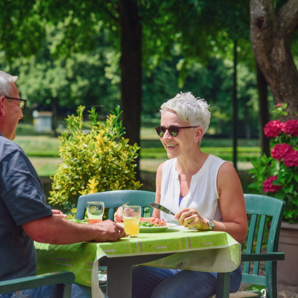 Zwei Personen sitzen an einem Tisch mit grünem Tischtuch im Freien, umgeben von Bäumen und blühenden Pflanzen.