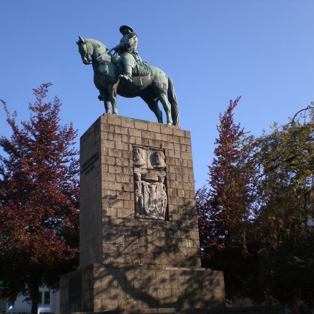 Der Große Kurfürst Friedrich Wilhelm als Bronzestatue auf einem Steinsockel. Er sitzt auf einem Pferd, trägt einen Hut und eine Uniform. Im Hintergrund Bäume.