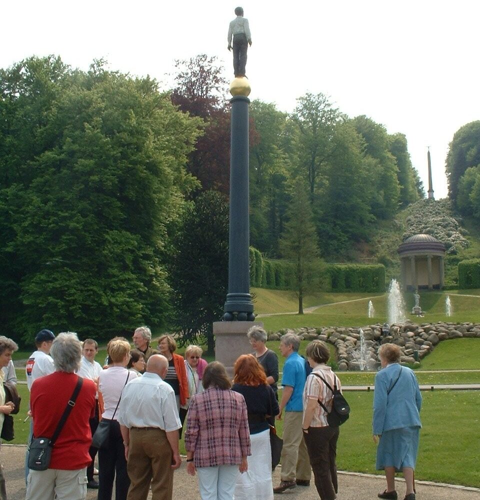 Gruppe von Menschen steht vor einer hohen Säule mit einer goldenen Kugel und einer Statue eines Mannes darauf, dem Eisernen Mann, umgeben von Park mit Bäumen, Brunnen und Pavillon.