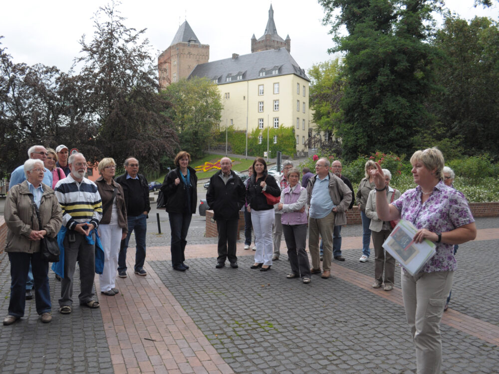 Eine Gruppe von Menschen steht im Freien auf gepflastertem Boden vor der Schwanenburg mit Türmen, eine Frau hält Papiere und spricht zur Gruppe.