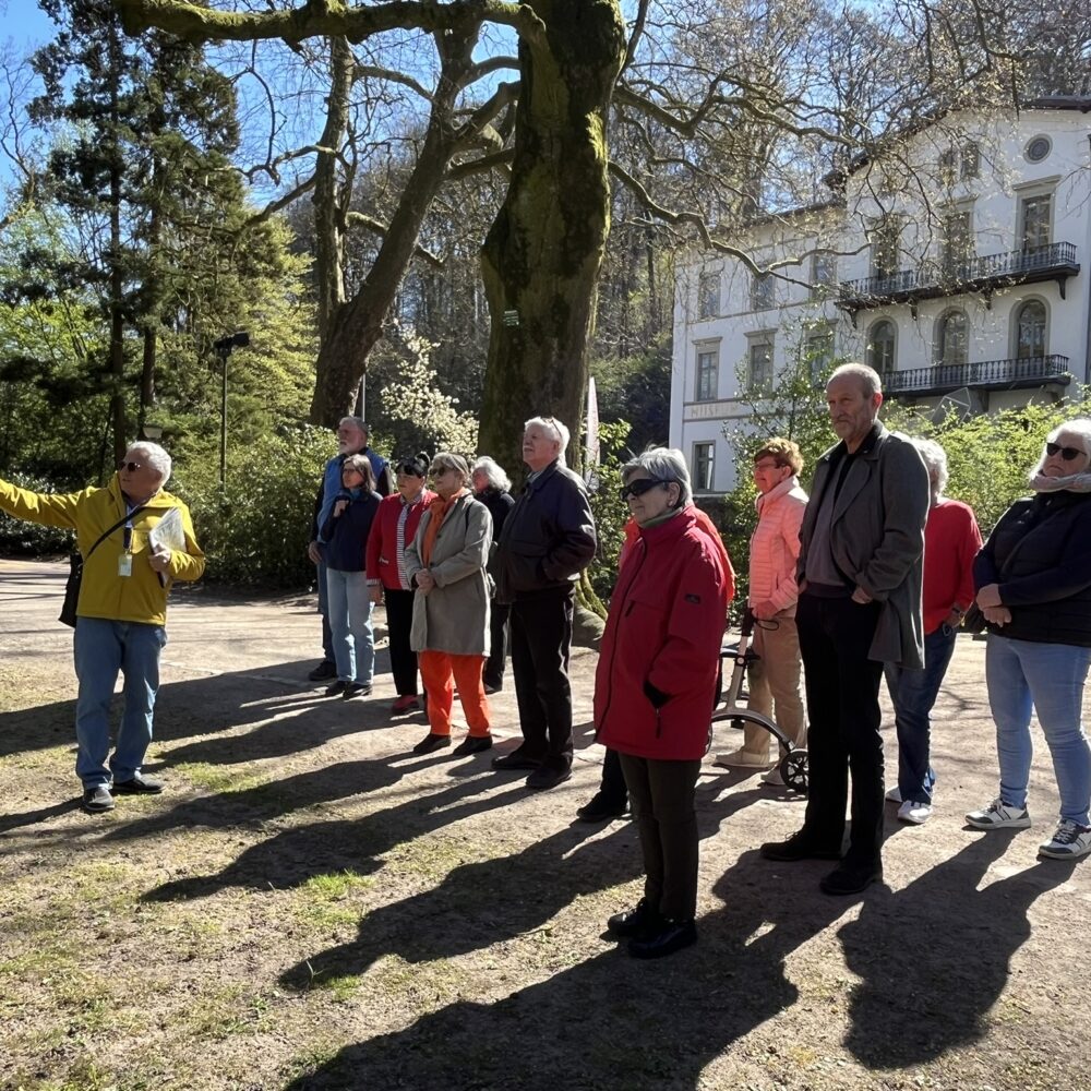 Eine Gruppe von Personen steht auf einem Weg in einem Park. Eine Person zeigt mit dem Finger in eine Richtung. Im Hintergrund ist ein Gebäude mit mehreren Fenstern und einem Balkon sichtbar.