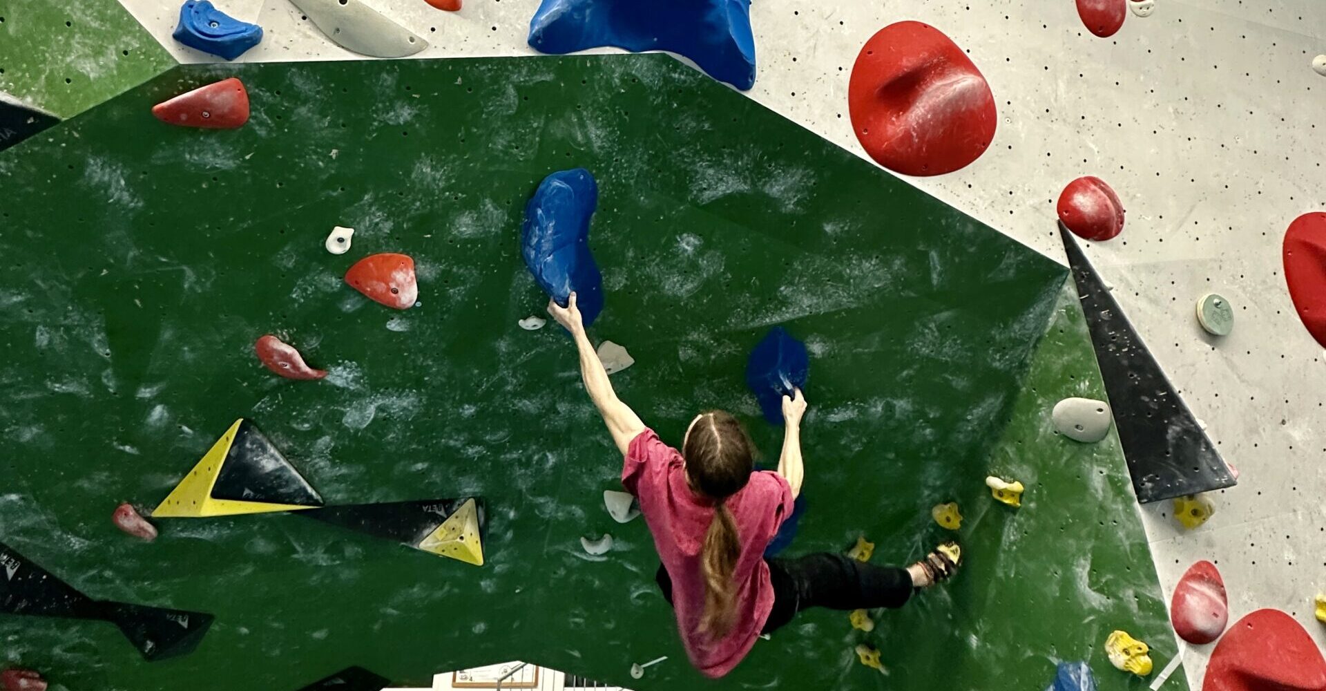 Eine Person mit langen Haaren in einem pinken Shirt klettert an einer Kletterwand mit grüner Basis und bunten Griffen in Rot, Blau und Gelb.
