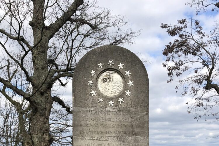 Denkmal für Johanna Sebus, grauer Stein mit einer runden Medaille und Sternen, umgeben von einer halbkreisförmigen Bank aus Beton, Bäume im Hintergrund.
