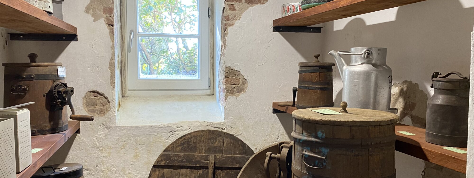 Kleiner Raum mit weiß verputzten Wänden, Fenster, Holzregalen und mehreren alten Holzfässern, Metallkannen und Käseformen auf den Regalen im Käsemuseum auf Gut Hogefeld