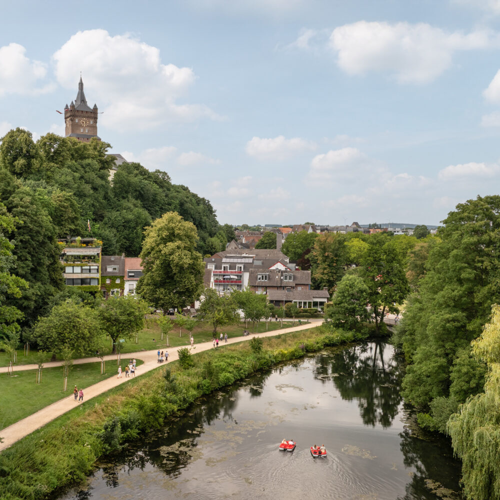 Blick auf einen Fluss mit drei Ruderbooten, umgeben von Bäumen und Häusern, im Hintergrund die Schwanenburg in Kleve auf einem bewaldeten Hügel unter bewölktem Himmel.