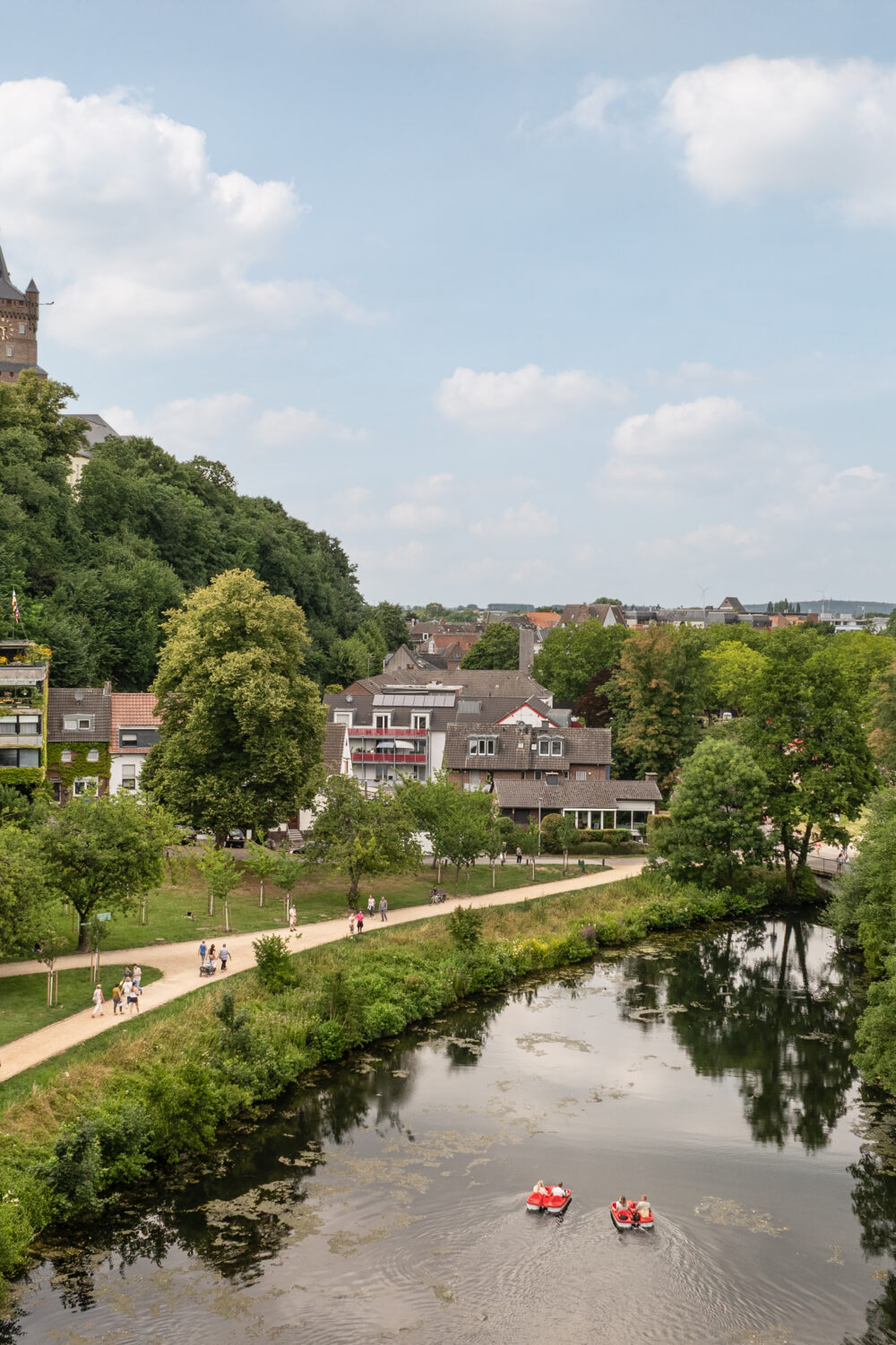 Blick auf einen Fluss mit drei Ruderbooten, umgeben von Bäumen und Häusern, im Hintergrund die Schwanenburg in Kleve auf einem bewaldeten Hügel unter bewölktem Himmel.