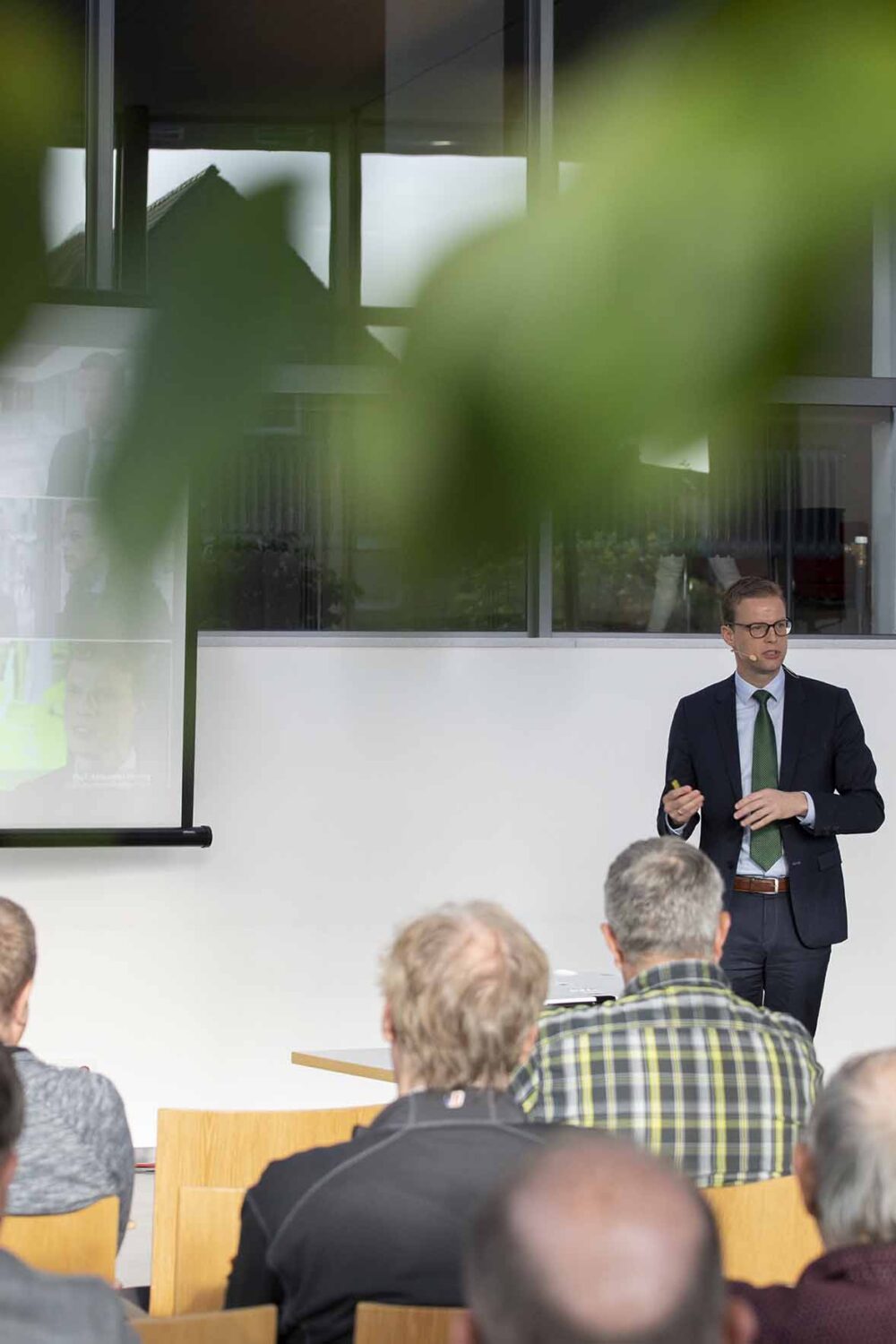 Mann im Anzug hält Vortrag vor Publikum im Foyer der Stadthalle Kleve, projizierte Bilder an der Wand, Blick durch grüne Blätter im Vordergrund