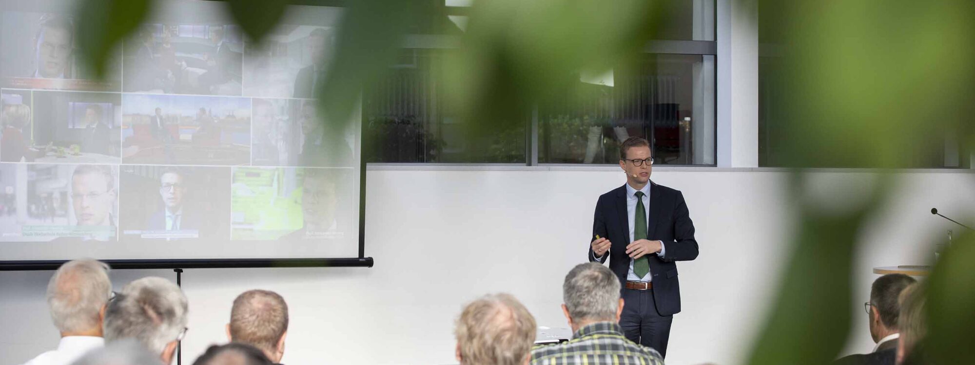 Mann im Anzug hält Vortrag vor Publikum im Foyer der Stadthalle Kleve, projizierte Bilder an der Wand, Blick durch grüne Blätter im Vordergrund
