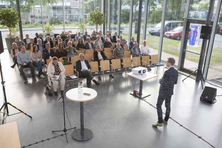 Ein Mann hält einen Vortrag vor einem Publikum in einem hellen Raum mit großen Fenstern in der Stadthalle Kleve und Blick auf eine Straße mit parkenden Autos.