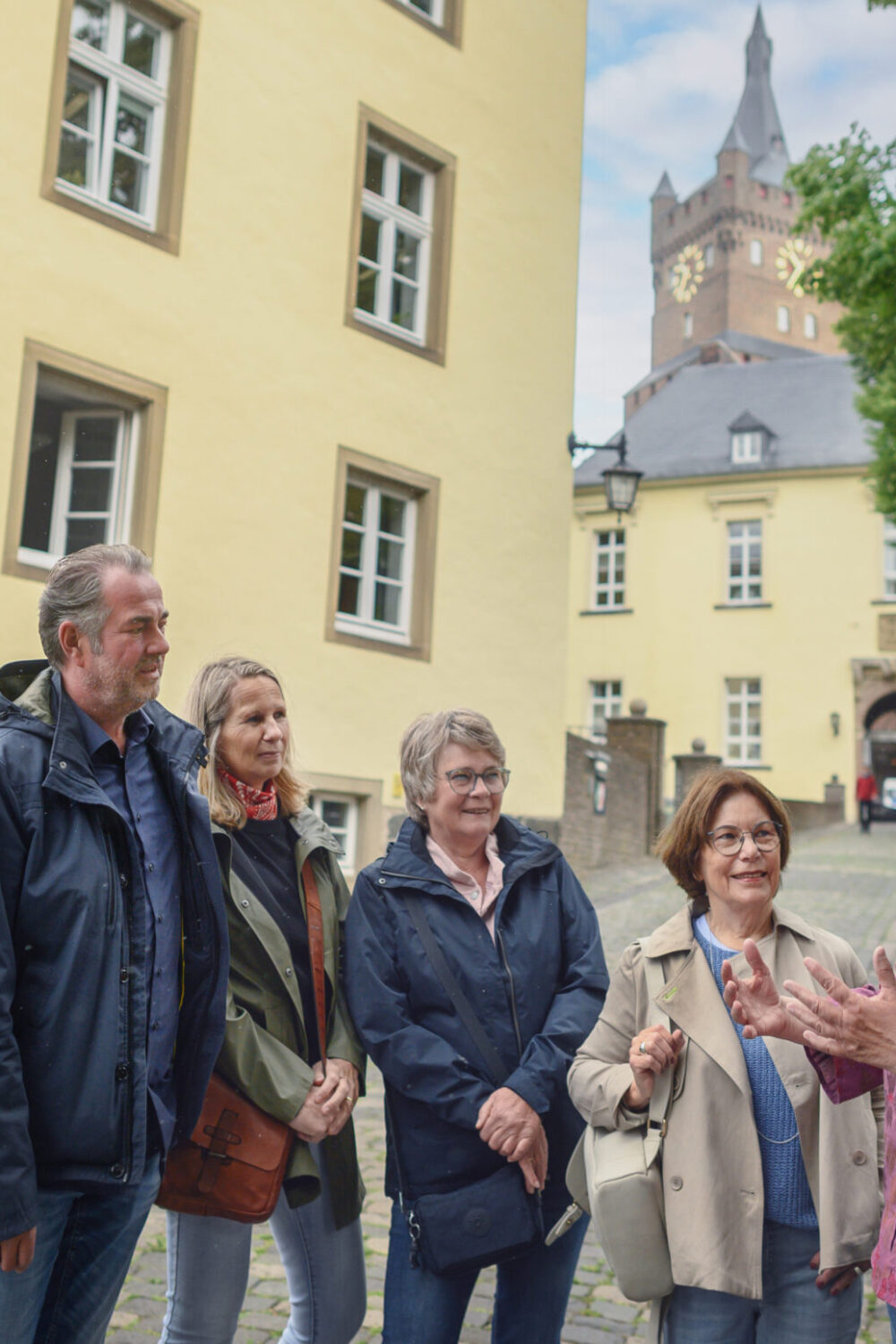 Fünf Personen stehen auf einem gepflasterten Weg vor einem gelben Gebäude mit einer Steinmauer. Im Hintergrund ist die Schwanenburg sichtbar. Zur erkennen ist eine Gästeführerin die Stadtführungen in Kleve anbietet.