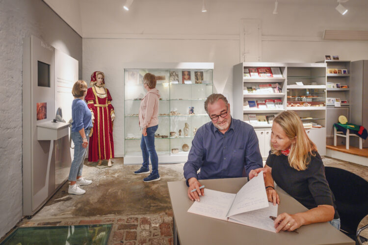 Drei Personen stehen in einem Ausstellungsraum, eine in einem historischen Kostüm. Ein Tisch mit Notizen und einem Laptop ist im Vordergrund.
