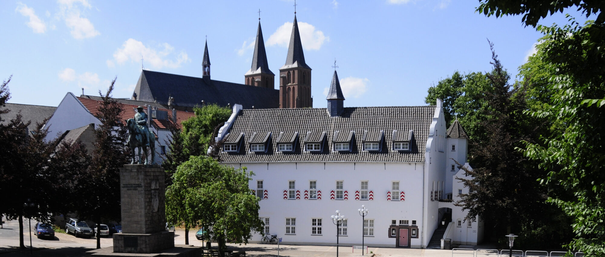 Blick auf das Marstallgebäude mit einem Reiterstandbild vom Großen Kurfürst im Vordergrund. Im Hintergrund sind zwei hohe Türme der Stiftskirche sichtbar. Das Gebäude hat ein Satteldach und Fenster mit roten und weißen Fahnen.