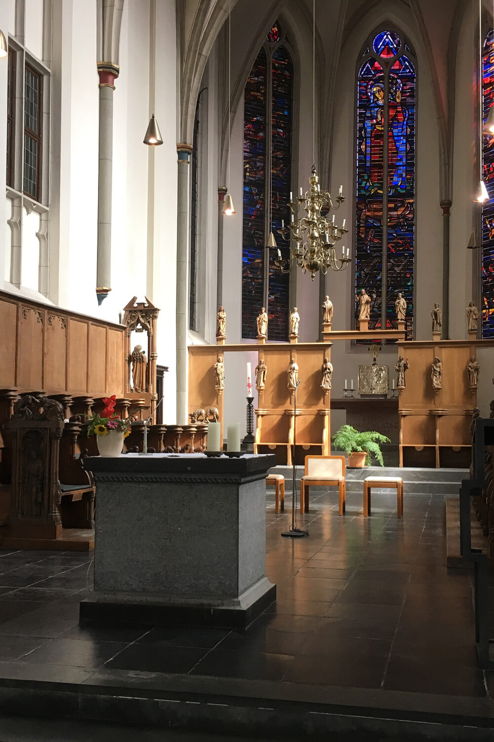 Innenansicht der Minoritenkirche mit einem Altar aus grauem Stein, Holzskulpturen an den Wänden, einer großen, bunten Glasfensterfront und einer Pflanzenarrangement auf einem Tisch.