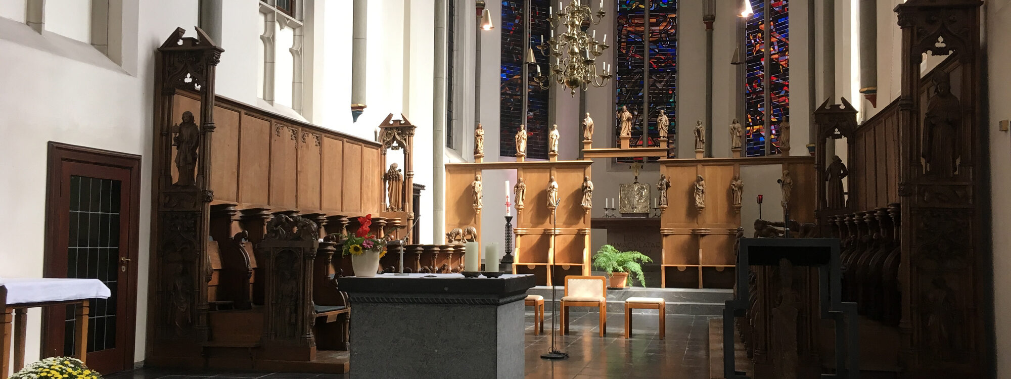 Innenansicht der Minoritenkirche mit einem Altar aus grauem Stein, Holzskulpturen an den Wänden, einer großen, bunten Glasfensterfront und einer Pflanzenarrangement auf einem Tisch.