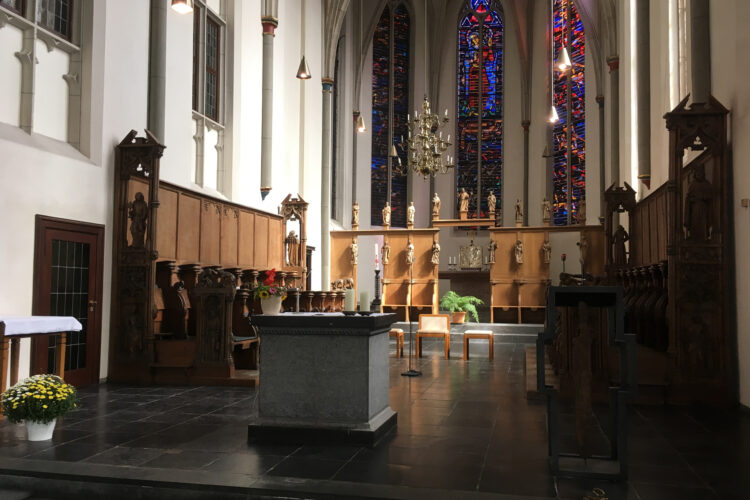 Innenansicht der Minoritenkirche mit einem Altar aus grauem Stein, Holzskulpturen an den Wänden, einer großen, bunten Glasfensterfront und einer Pflanzenarrangement auf einem Tisch.