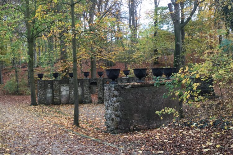 Zwei alte, teilweise verfallene Mauern mit mehreren schwarzen Behältern auf der Oberseite, umgeben von herbstlichen Bäumen mit buntem Laub.