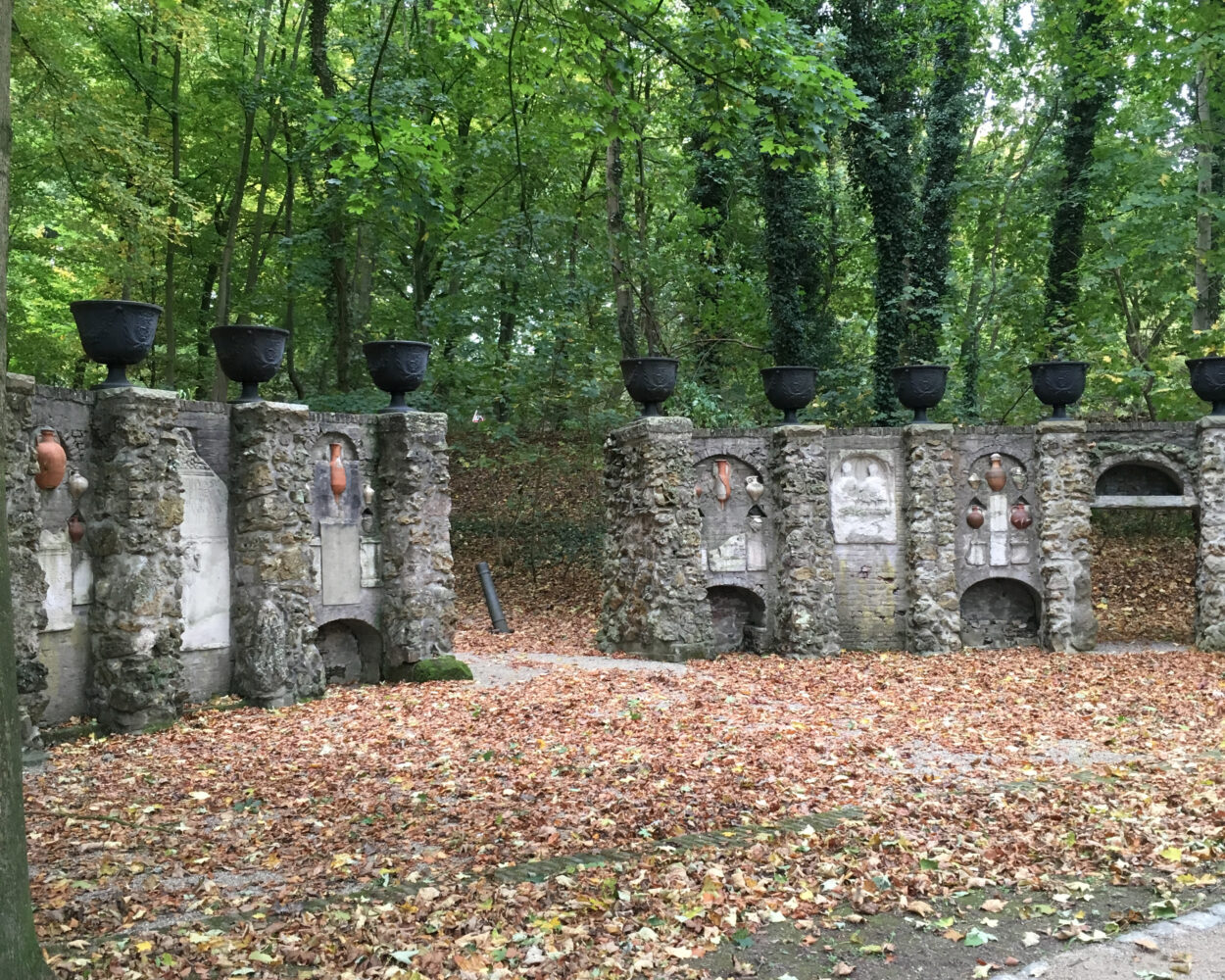 Zwei alte, teilweise verfallene Mauern mit mehreren schwarzen Behältern auf der Oberseite, umgeben von herbstlichen Bäumen mit buntem Laub.