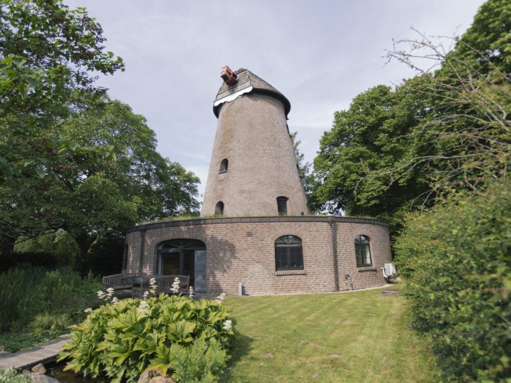 Runde Mühle mit grauem Turm und Backsteinsockel, umgeben von grünem Garten und Bäumen unter blauem Himmel.