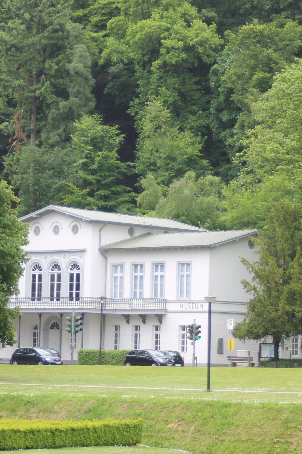 Weißes Gebäude des Museums Kurhaus Kleve mit mehreren Fenstern, umgeben von grünen Bäumen und Rasenflächen der historischen Gartenanlagen.