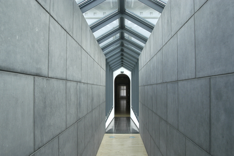 Langer, schmaler Gang mit grauen Betonwänden im Museum Kurhaus Kleve und einem Glasdach, das Licht hereinlässt, am Ende ein dunkler Durchgang.