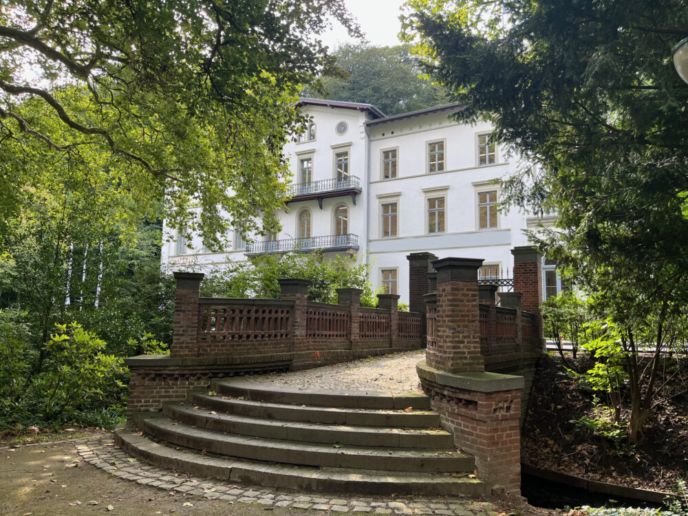 Weißes dreistöckiges Gebäude des Museum Kurhaus Kleve mit Balkonen, umgeben von Bäumen, davor eine breite Treppe mit Brücke und Ziegelgeländer.