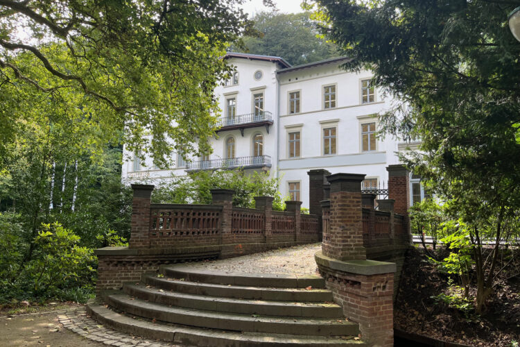 Weißes dreistöckiges Gebäude des Museum Kurhaus Kleve mit Balkonen, umgeben von Bäumen, davor eine breite Treppe mit Brücke und Ziegelgeländer.