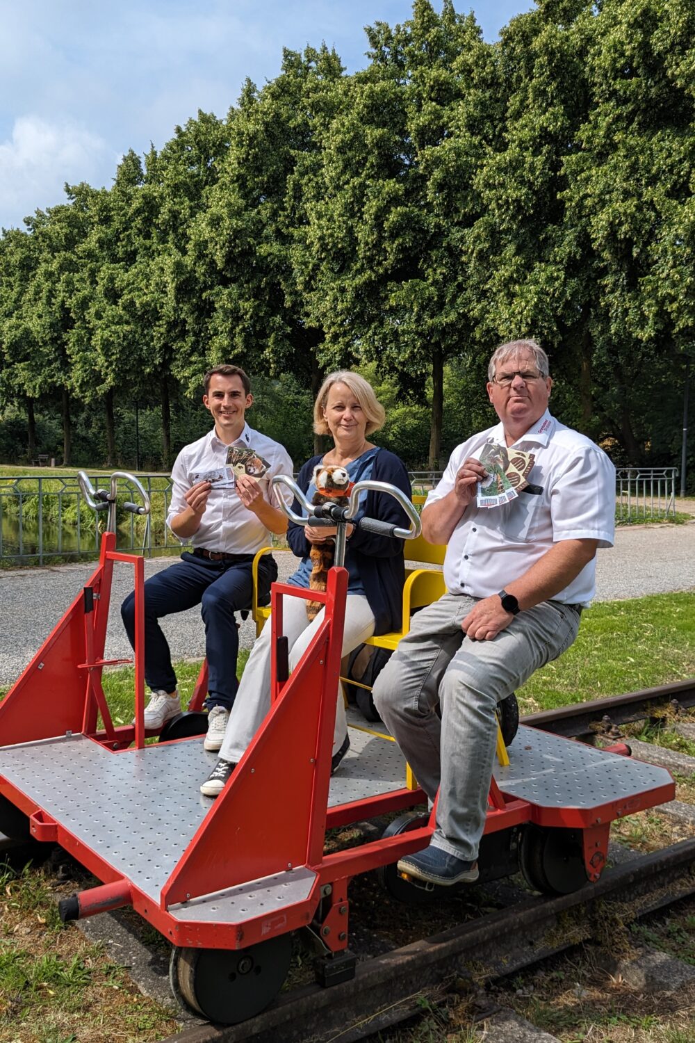 Drei Personen sitzen an einem gelben Tisch auf einer Draisine. Sie halten Gläser in der Hand und stoßen an. Die Draisine hat rote Geländer.