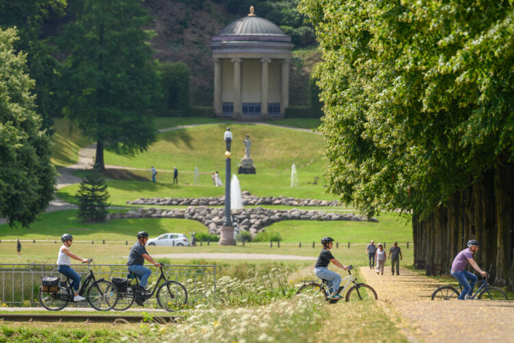 Vier Radfahrer fahren auf einem von Bäumen gesäumten Weg in einem Park, im Hintergrund ein klassizistischer Pavillon mit Säulen und Kuppeldach.