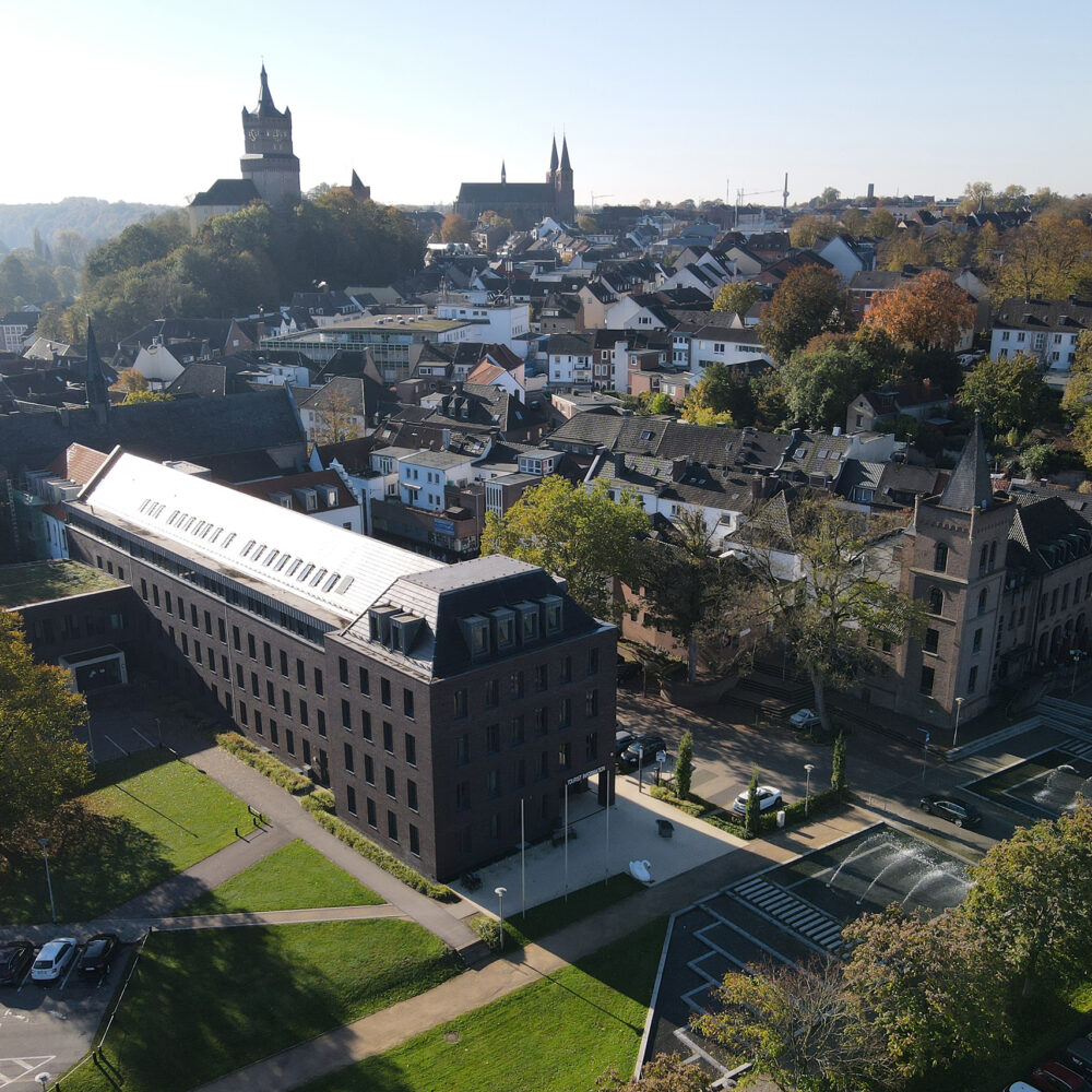 Luftaufnahme von Kleve mit dem modernen Rathausgebäude im Vordergrund inklusive der Tourist Information und der Schwanenburg auf einem Hügel im Hintergrund, umgeben von Wohnhäusern und Bäumen.