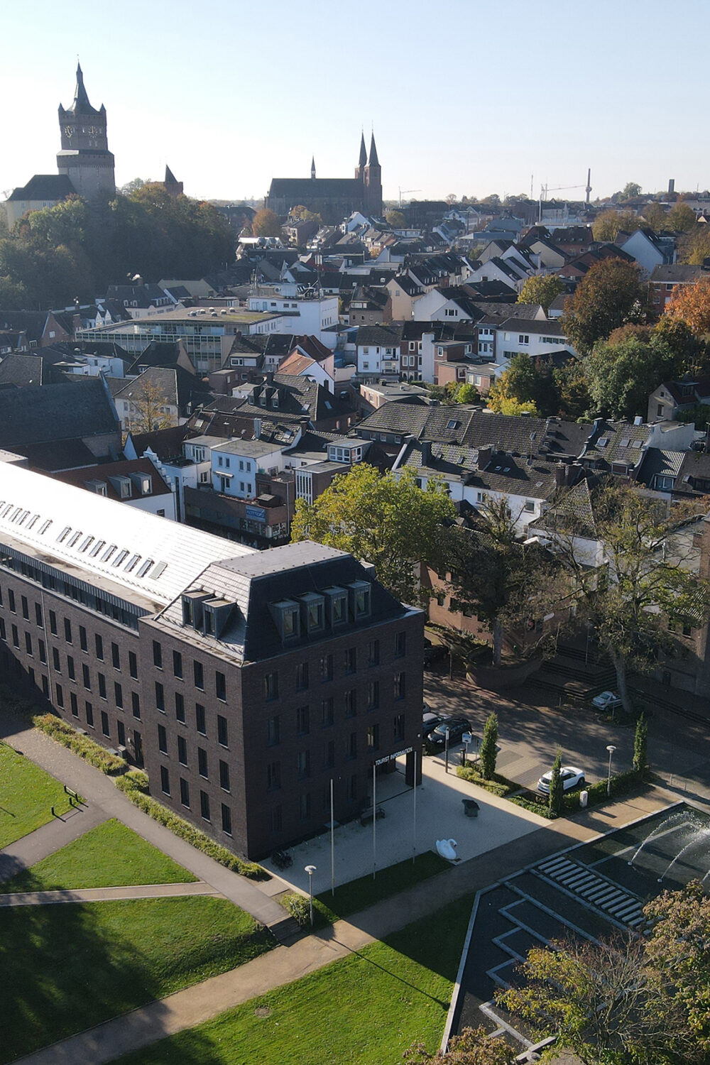 Luftaufnahme von Kleve mit dem modernen Rathausgebäude im Vordergrund inklusive der Tourist Information und der Schwanenburg auf einem Hügel im Hintergrund, umgeben von Wohnhäusern und Bäumen.