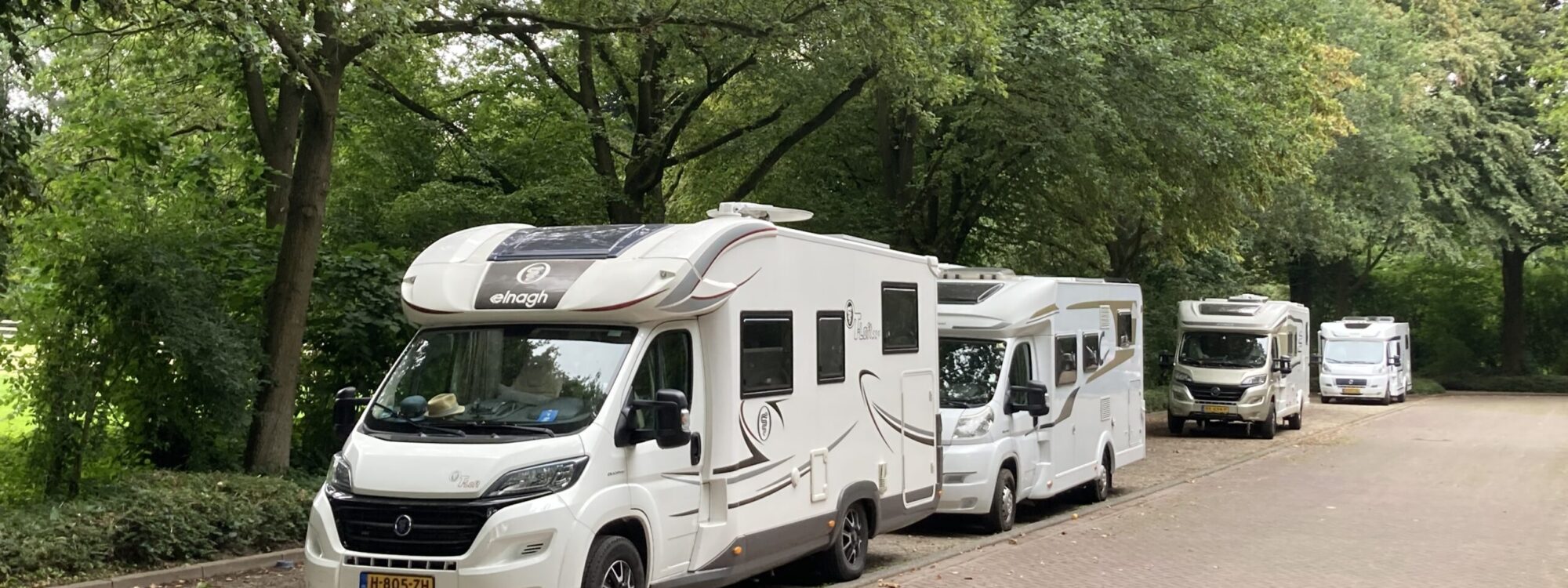 Vier weiße Reisemobile stehen hintereinander auf einem gepflasterten Stellplatz unter grünen Bäumen.