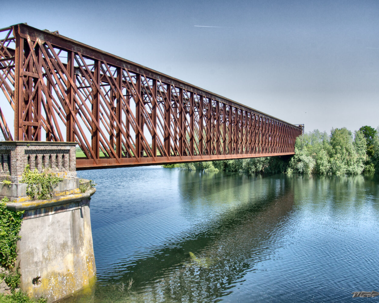 Eisenbahnbrücke mit rotem Stahlträgerwerk über einem ruhigen Fluss, umgeben von Bäumen und Sträuchern. Wasser spiegelt die Brücke wider.