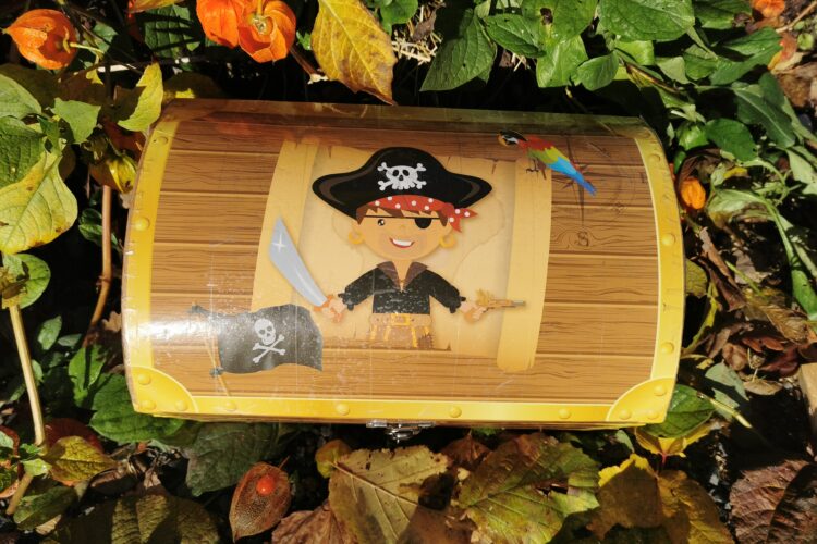 Eine Schatzkiste mit einem Piratenmotiv, das einen Piraten mit Schwert und Pistole zeigt. Die Kiste hat eine Holzoptik und ist mit einem Totenkopf-Symbol verziert.