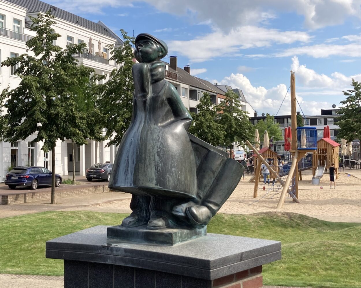 Bronzeskulptur des Schüsterken, einer Figur in traditioneller Kleidung mit einem großen Hut und einem Umhang, steht auf einem Sockel. Im Hintergrund sind Spielgeräte und Wohngebäude sichtbar.