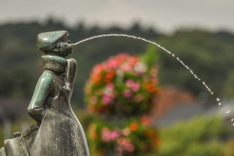 Bronze-Skulptur eines Jungen mit einer Mütze, der Wasser aus dem Mund spritzt. Im Hintergrund blühende Pflanzen und unscharfe Landschaft.