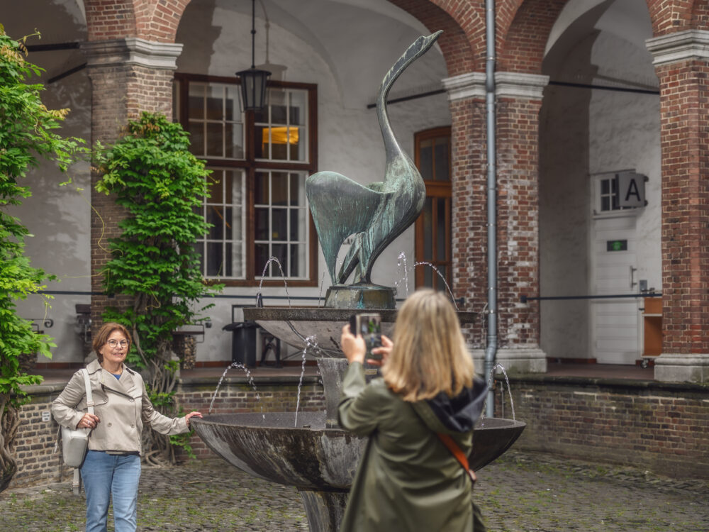 Bronzeskulptur eines Schwans auf einem Brunnen in einem Innenhof, umgeben von Pflanzen und historischen Gebäuden. Sichtbare Person fotografiert die Skulptur.